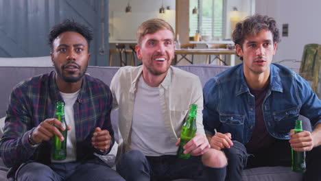 multi-cultural group of male friends watching sports game on tv at home celebrating goal with beer