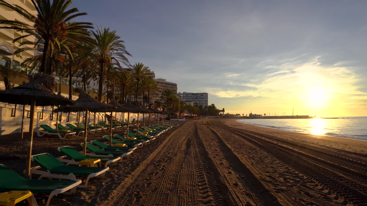 POV camminando lungo la spiaggia di Marbella all'alba con gimbal, lato sinistro con lettini e ombrelloni con palme tropicali, lato destro mare Mediterraneo, ispirazione per viaggi di lusso