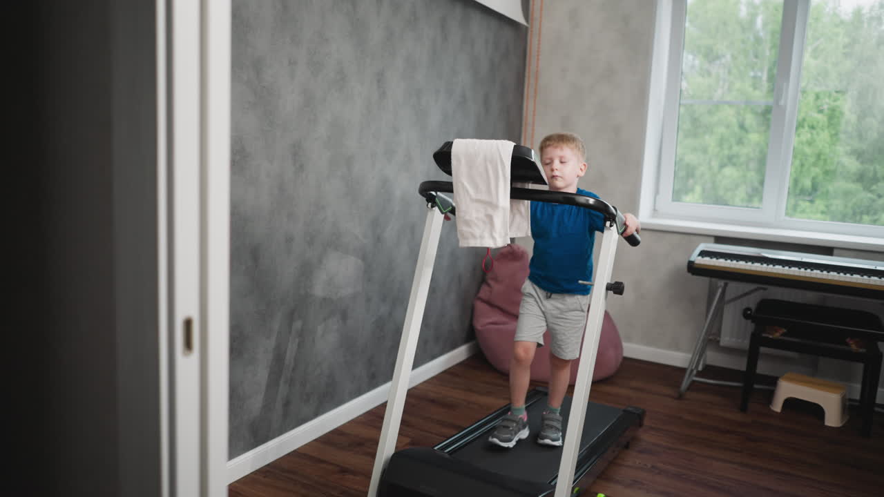 Curious kid walking on treadmill indoors, holding front handle with towel draped over bar, gazing toward background window with focused expression, modern room with piano and wooden floor visible