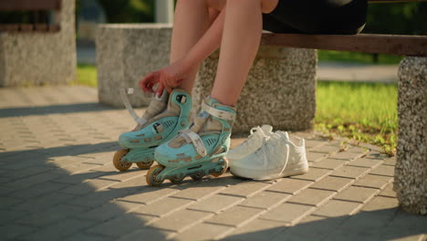 lower view of individual adjusting roller skate on right leg while seated on bench outdoors, with white sneaker placed behind, sunlight casting warm shadows, greenery visible in background