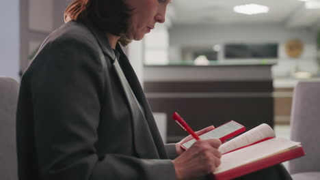 businesswoman sitting in modern office, looking at red notebook and mobile phone, multitasking, focused on task, reading, corporate workspace, gray suit, productivity, concentration