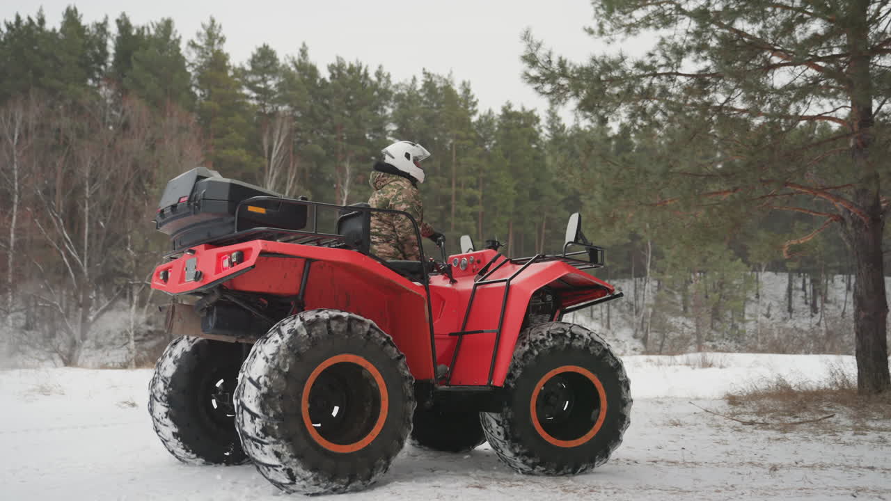 Un pilota di quad rosso in una foresta innevata, conducente con casco e giacca mimetica seduto su un potente quad rosso con pneumatici sovradimensionati, baule posteriore sul portapacchi, pattugliamento lento lungo un sentiero costeggiato da pini, tracce fresche nella neve soffice.