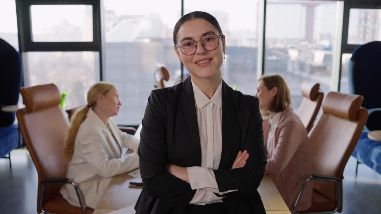 Portrait of a confident brunette businesswoman in round glasses in a black business suit who folds her arms on her chest and poses confidently in a modern office with large windows