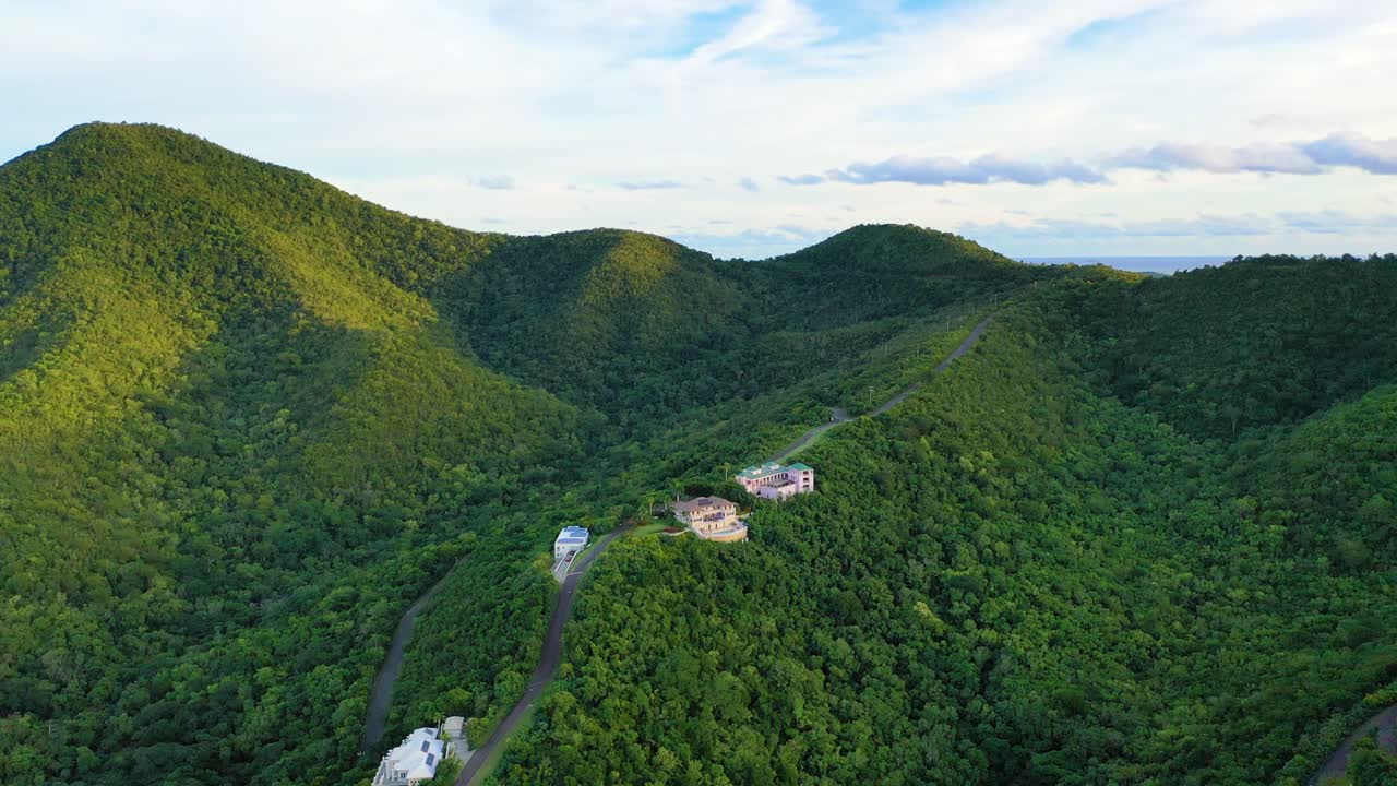 Aerial drone footage over St. Croix reveals a few luxury homes nestled in a dense forest, surrounded by lush rolling hills and island mountain ridges glowing under bright midday Caribbean sun
