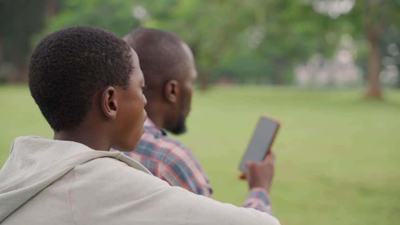 Black father and son sharing phone on lawn, father demonstrates screen gesture while son watches and asks questions, casual hoodie and plaid shirt, modern parenting scene in green park, learning