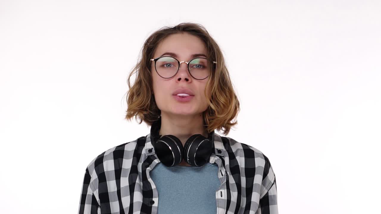 Attractive young woman with hedphones on neck blowing chewing gum bubble with eyes wide open isolated over white background. Chewing gum bursts on the chin and she's starting to laugh. Slow motion