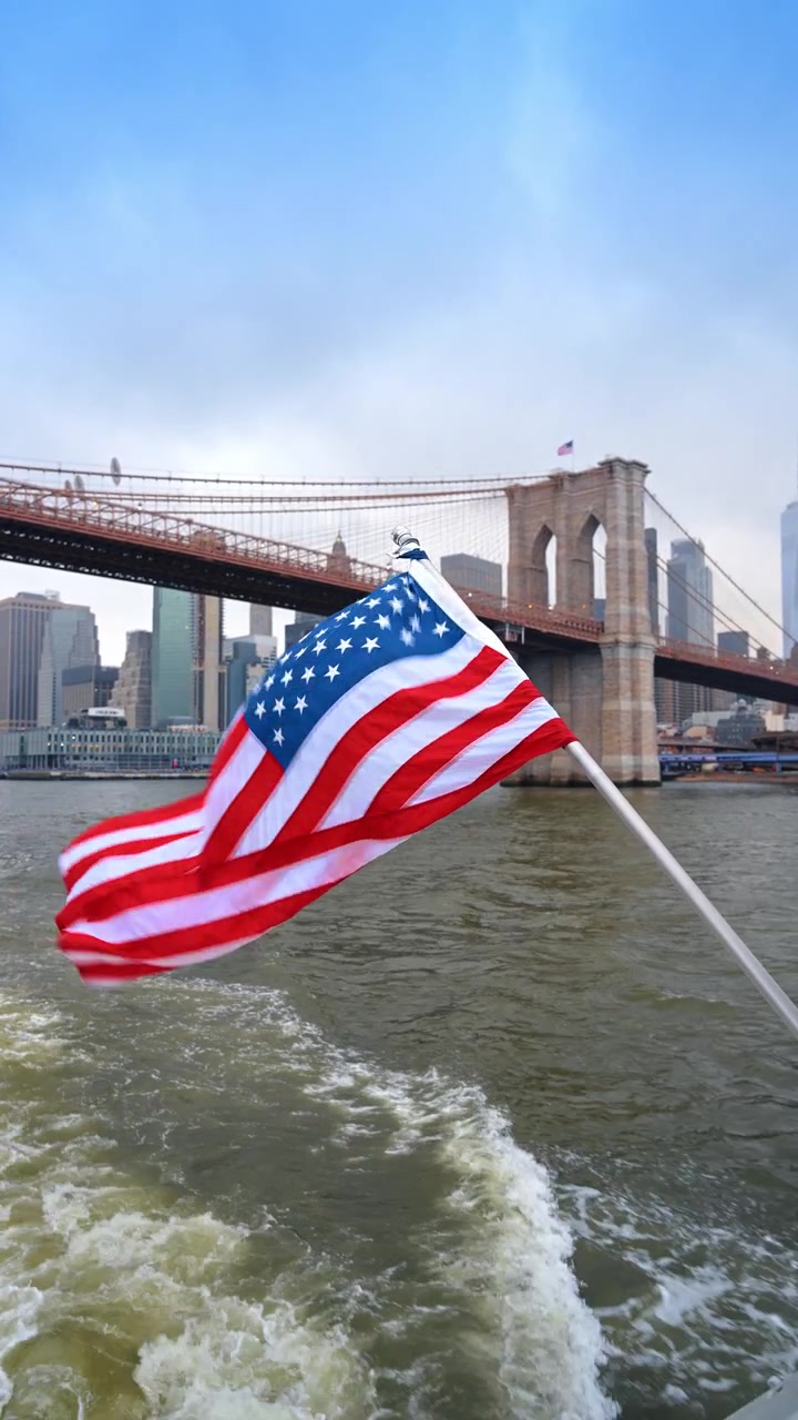 Bandiera americana che sventola vicino al Ponte di Brooklyn, New York. Scia di barca sull'East River con lo skyline di Manhattan e una torre di pietra sotto un cielo coperto