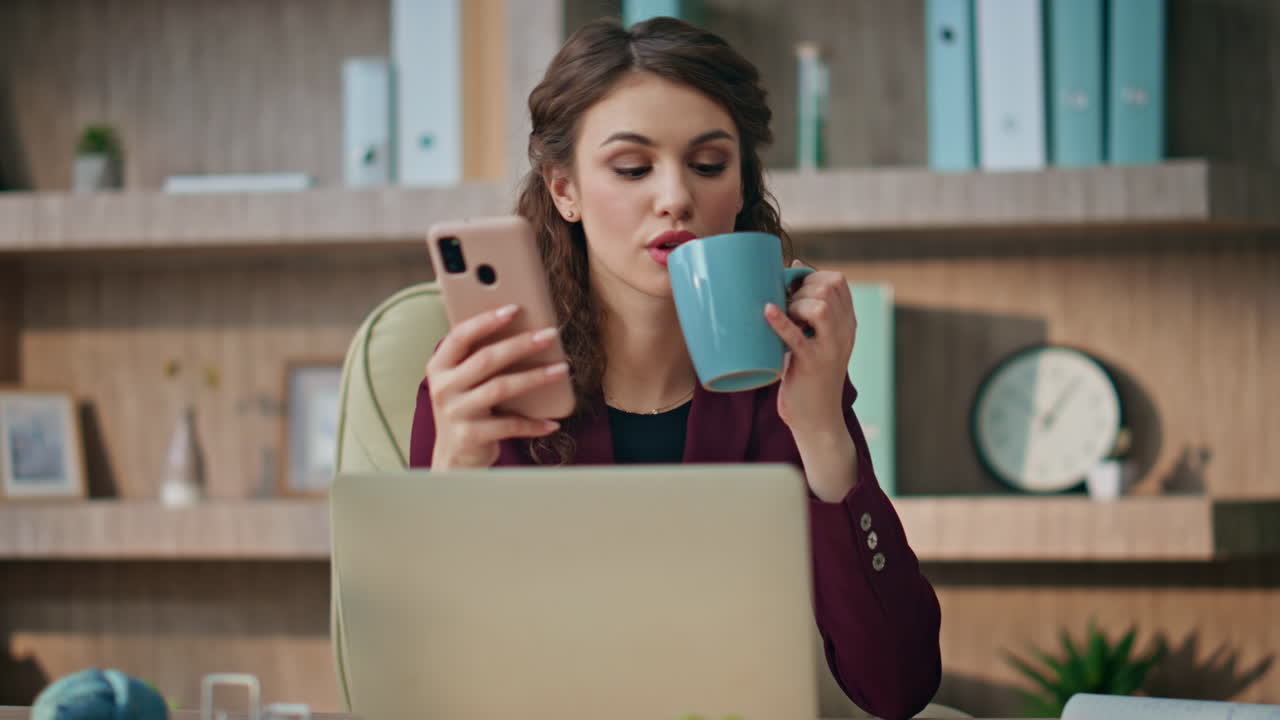 Morning businesswoman looking cellphone in office room. Bored girl drink coffee