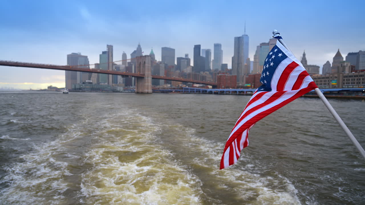 Bandiera statunitense su traghetto con skyline del porto di New York. Bandiera americana su un'imbarcazione in movimento con lo skyline di Lower Manhattan e il Ponte di Brooklyn sotto un cielo coperto.