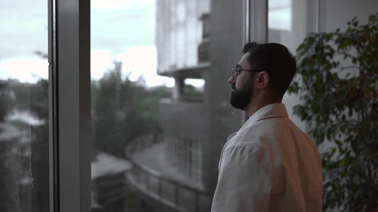 man with beard gazing out modern office building interior windows, thoughtful and contemplative side view, white green plants, glass concrete materials, business professional context.