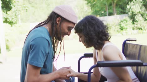 Biracial-couple-touching-heads-holding-hands-in-garden,-woman-in-wheelchair,-man-with-dreadlocks