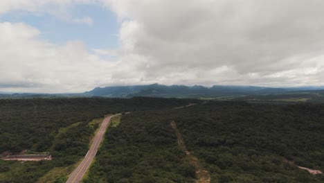 Road-through-green-forests,-mountains-and-clouds-in-the-distance,-panoramic-drone-view