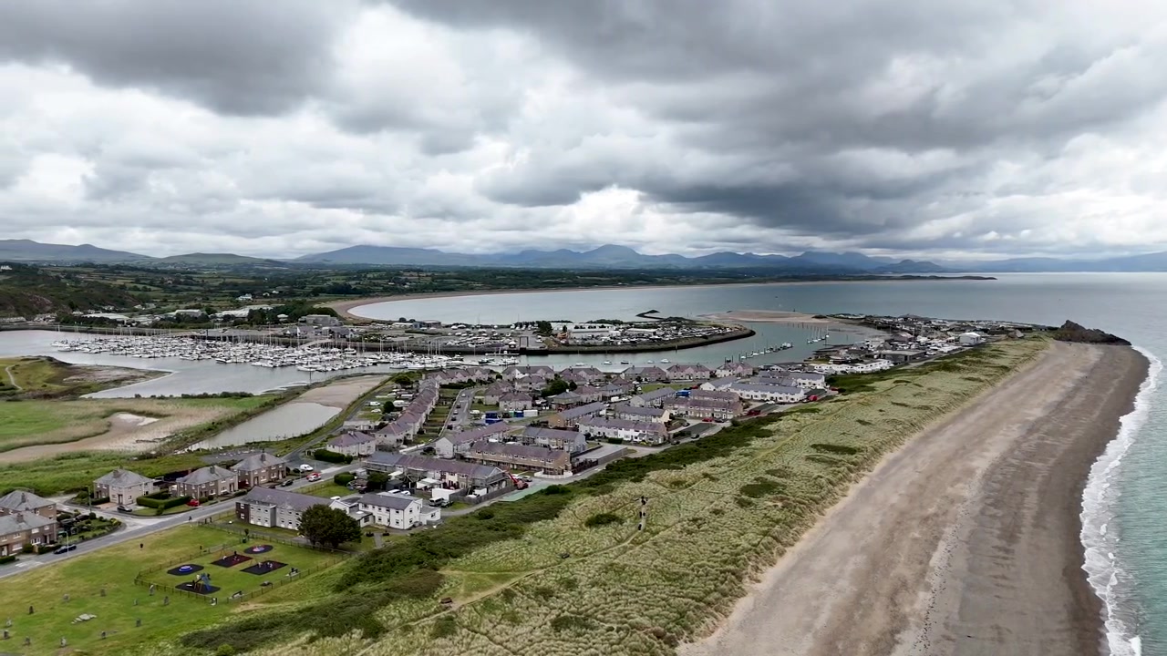 Vista aerea fluida con drone di un porto turistico costiero, una spiaggia sabbiosa e una città di mare lungo la costa del Galles del Nord. Costa panoramica con nuvole drammatiche e mare calmo catturata in 4K cinematografico