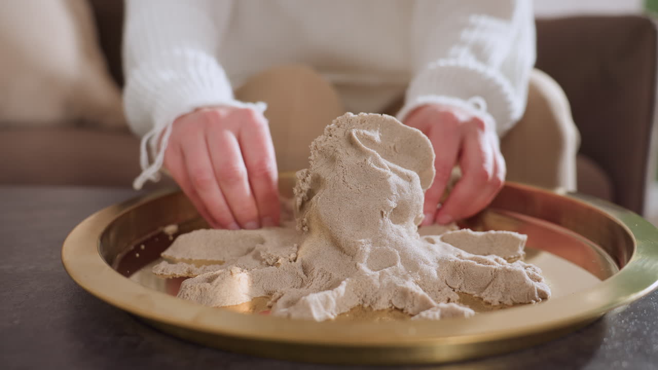 Lower view of lady hands delicately shaping octopus figure in soft soil mold on golden tray during creative therapy session, capturing tactile engagement and artistic focus in peaceful indoor setting