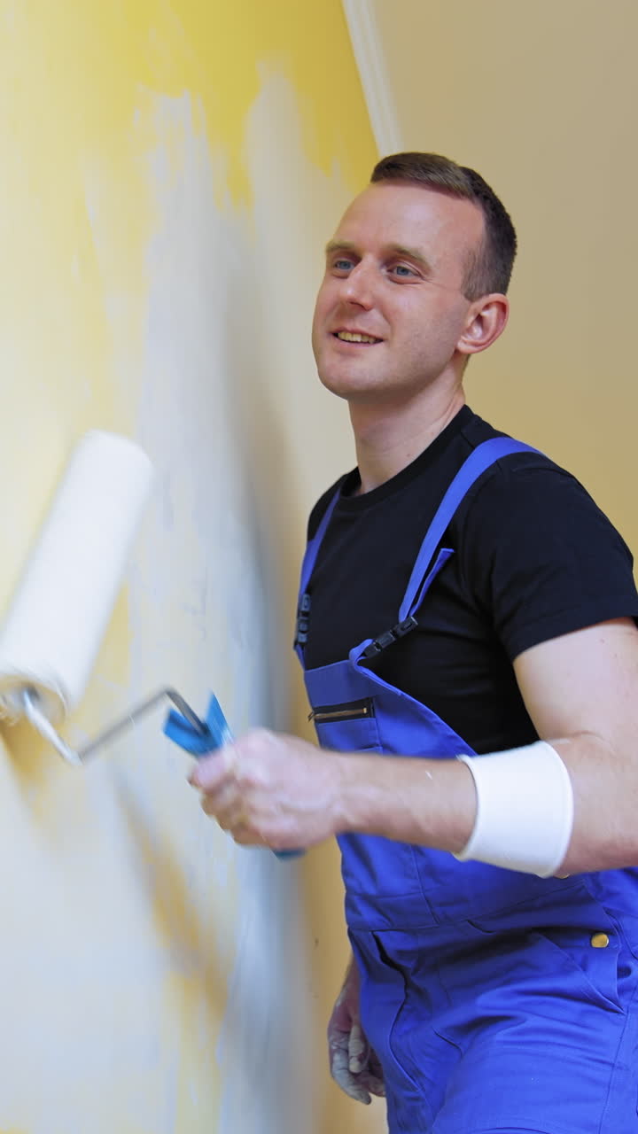 Young man improving apartment. Interior decoration in flat. Worker in blue overalls painting wall with a paint roller. Painting services. Vertical video