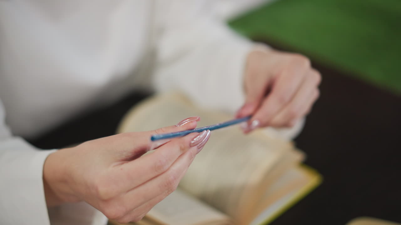 푸른 을 들고 있는 손의 클로즈업, 배경에 흐릿한 열린 책, 편안하고 지적 분위기를 만들고, 매니큐어 손과 현대적인 실내 환경을 특징으로합니다.