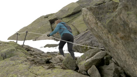 a woman hiking up steep, rocky and snowy terrain using a rope on an overcast day in norway, slow motion