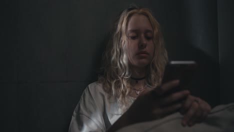 a young blonde girl in a white nightie scrolls social networks while lying in a gray bed near the wall of the house at night