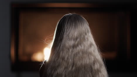 a young girl with long hair watches the fire burn in the fireplace