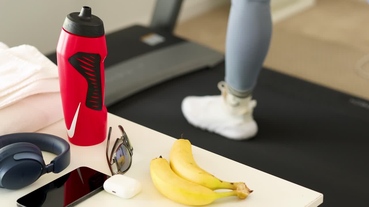Person walking on treadmill in gym setting, surrounded by fitness gear including water bottle and headphones