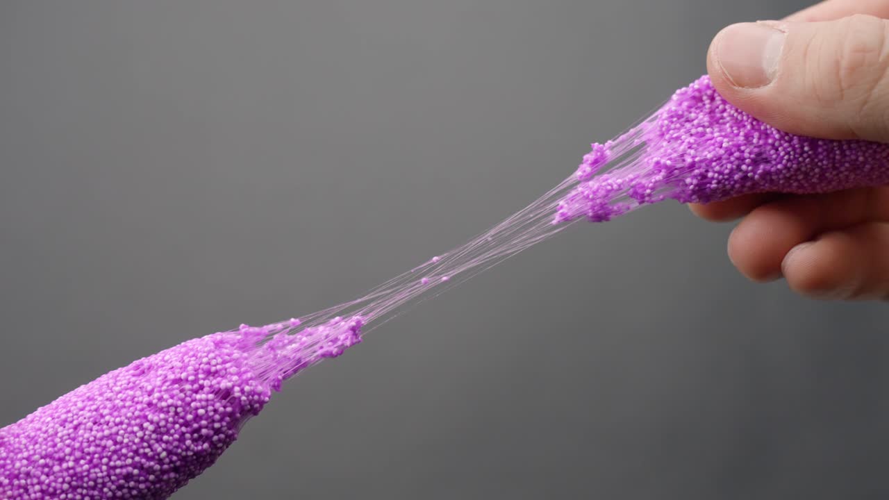 Two hands stretch and squeeze lilac floam slime with microbeads, revealing sticky threads and elastic movement in ASMR toy manipulation, gray background, static camera, real time, close-up framing