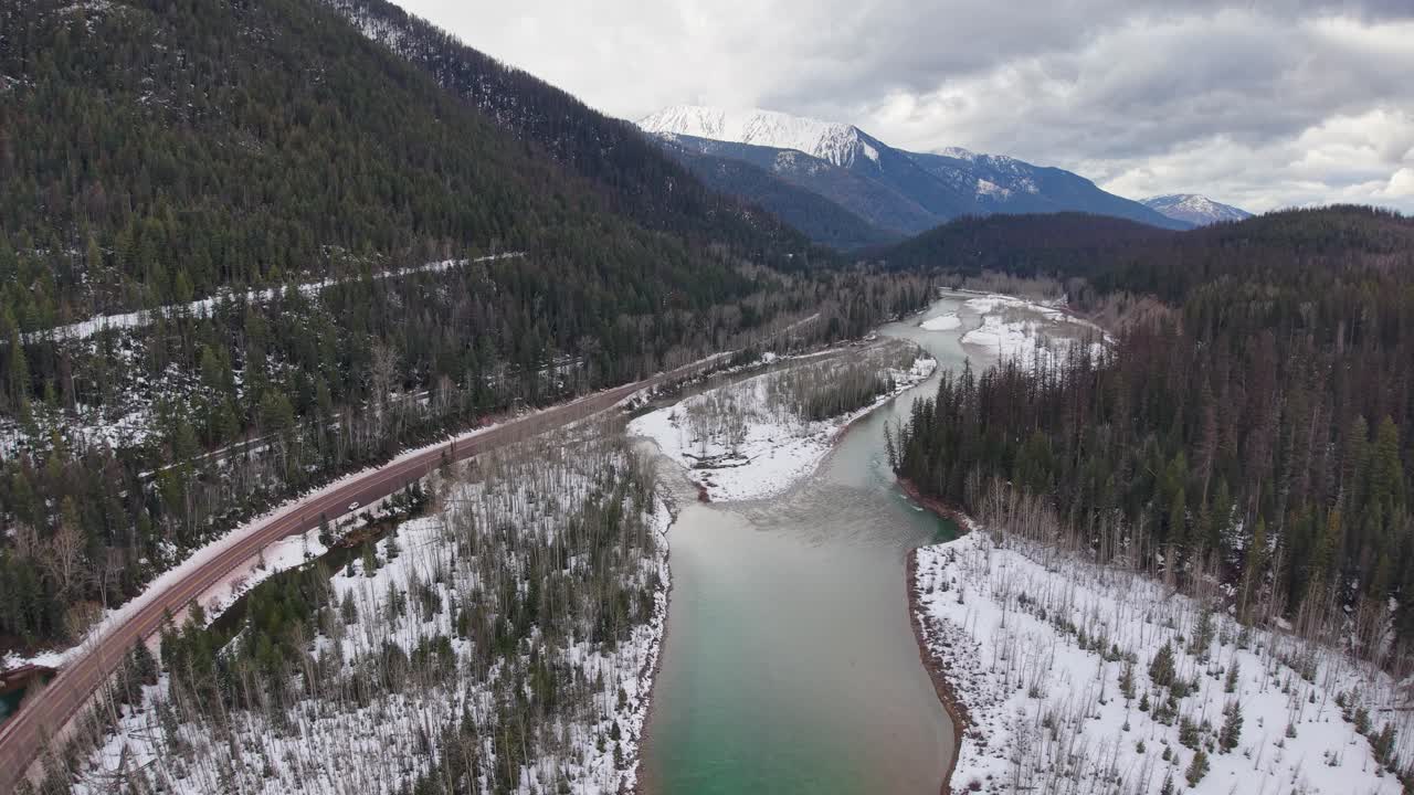 Riprese aeree con drone di un fiume serpeggiante che scorre lungo una strada forestale attraverso una valle imbiancata dalla neve, con colline sempreverdi e montagne distanti nel Parco Nazionale del Glacier, Montana.
