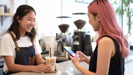 a young woman with pink hair interacts with a barista at a café, captivated by her smartphone as they share a friendly moment over iced coffee.