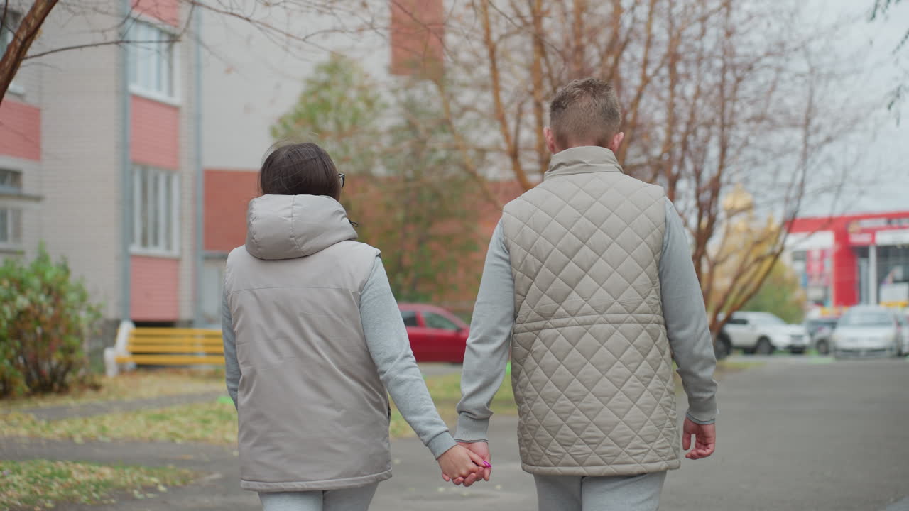 Back view of couple walking hand in hand during morning stroll wearing matching light-colored outfits, on park pathway with trees on both sides and colorful distant buildings in background