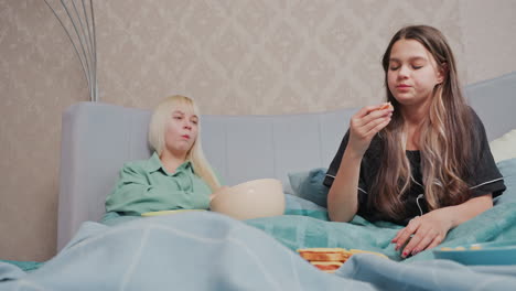 two young girls sit comfortably on bed sharing breakfast and laughter, one girl in black pajamas nods while picking up sandwich, other girl eats chips and smiles