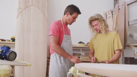 Two-Caucasian-male-surfboard-makers-discussing-the-project-with-surfboards-in-the-background