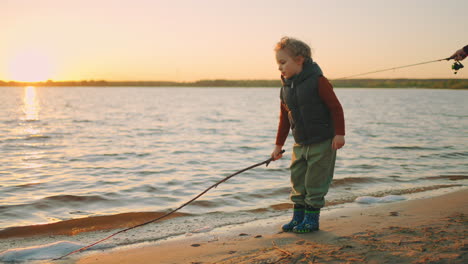 little curly boy is playing with stick on river shore touching water resting with his father fisher