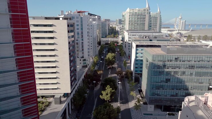 Avenida urbana com edifícios modernos em uma área de escritórios em Lisboa, com a Ponte Vasco da Gama ao fundo, em um plano de recuo.