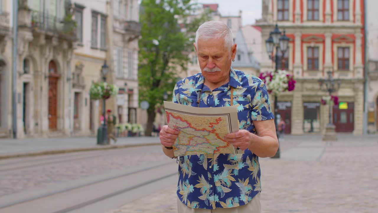 Anziano nonno turista elegante che cammina per strada cercando la via usando una mappa cartacea in città.