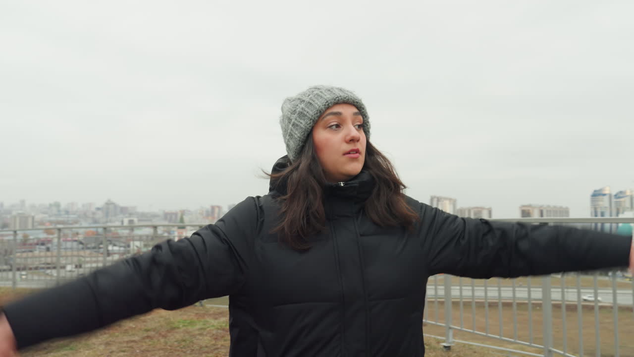 Danser spejler byen. En levende kvinde udfører en dans ved et gelænder under en panoramisk udsigt over byen. En ung kvindelig danser træner nær et gelænder med en omfattende byskyline i baggrunden.