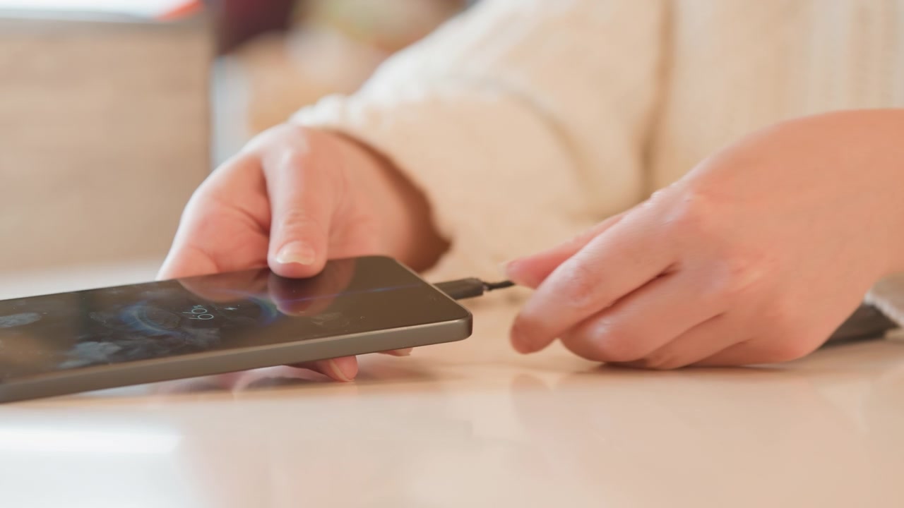 Close-up view of two mobile devices on tabletop with frayed charging cable linking ports, exposed inner wires, worn sheath, and hands guiding plug into place during careful troubleshooting