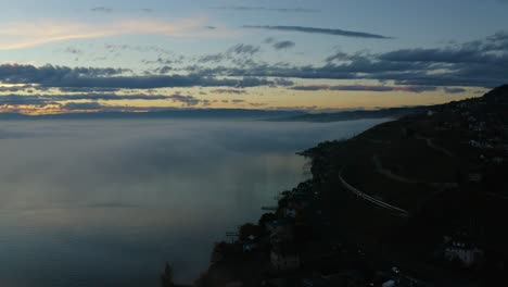 Überfliegendes-Ufer-Des-Genfersees-Mit-Vorbeifahrendem-Schweizer-Zug-Und-Verschwinden-Im-Nebel-Bei-Sonnenuntergang