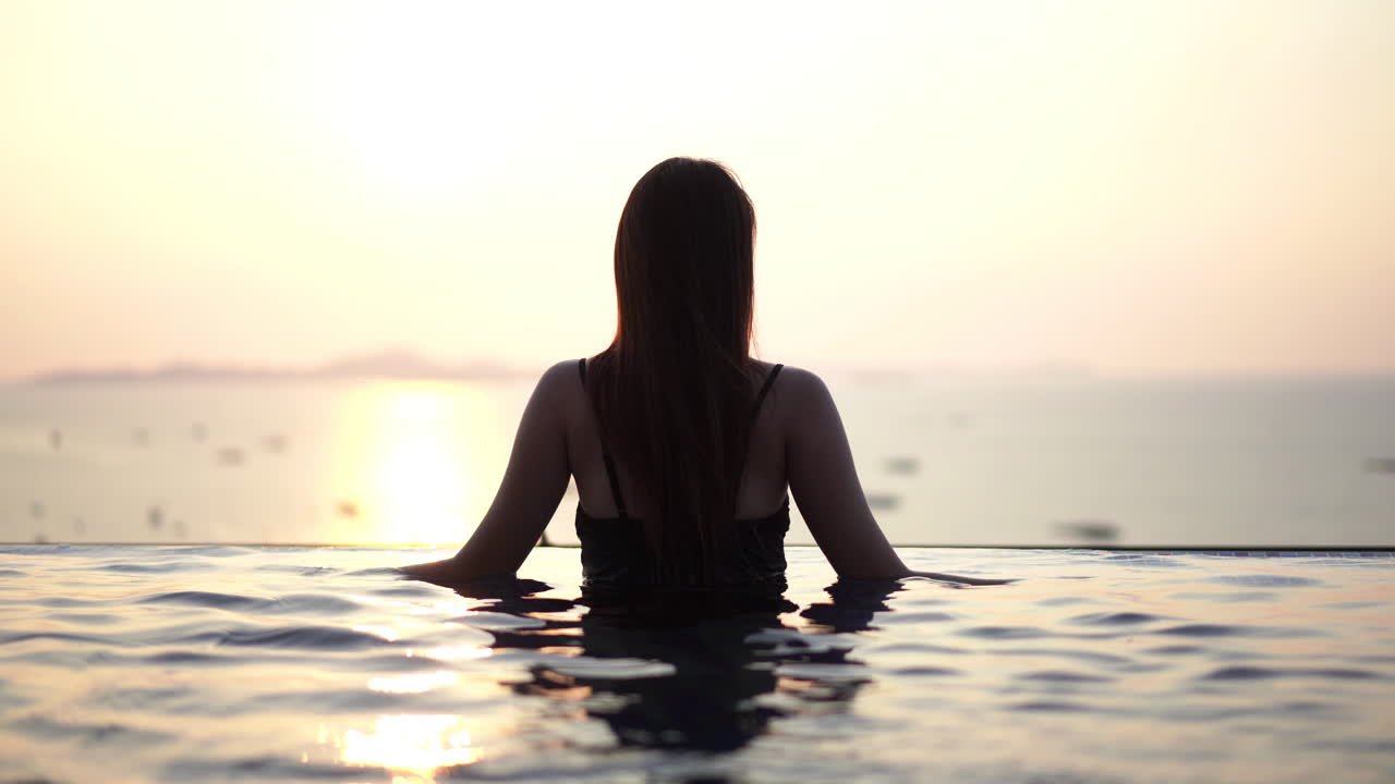 Silhouette einer Frau in einem Infinity-Pool mit Blick auf Boote im Meer bei Sonnenuntergang
