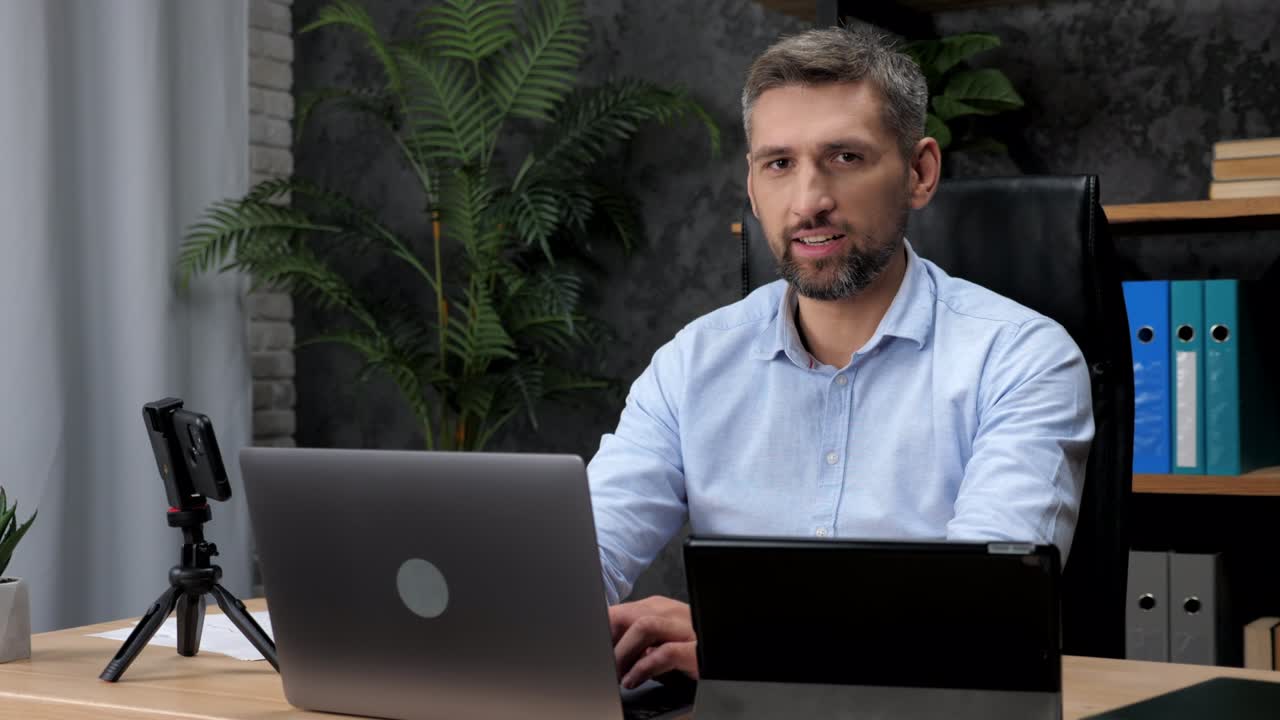 un hombre de negocios adulto trabaja para una computadora portátil hablando en línea se vuelve hacia la cámara y sonríe.