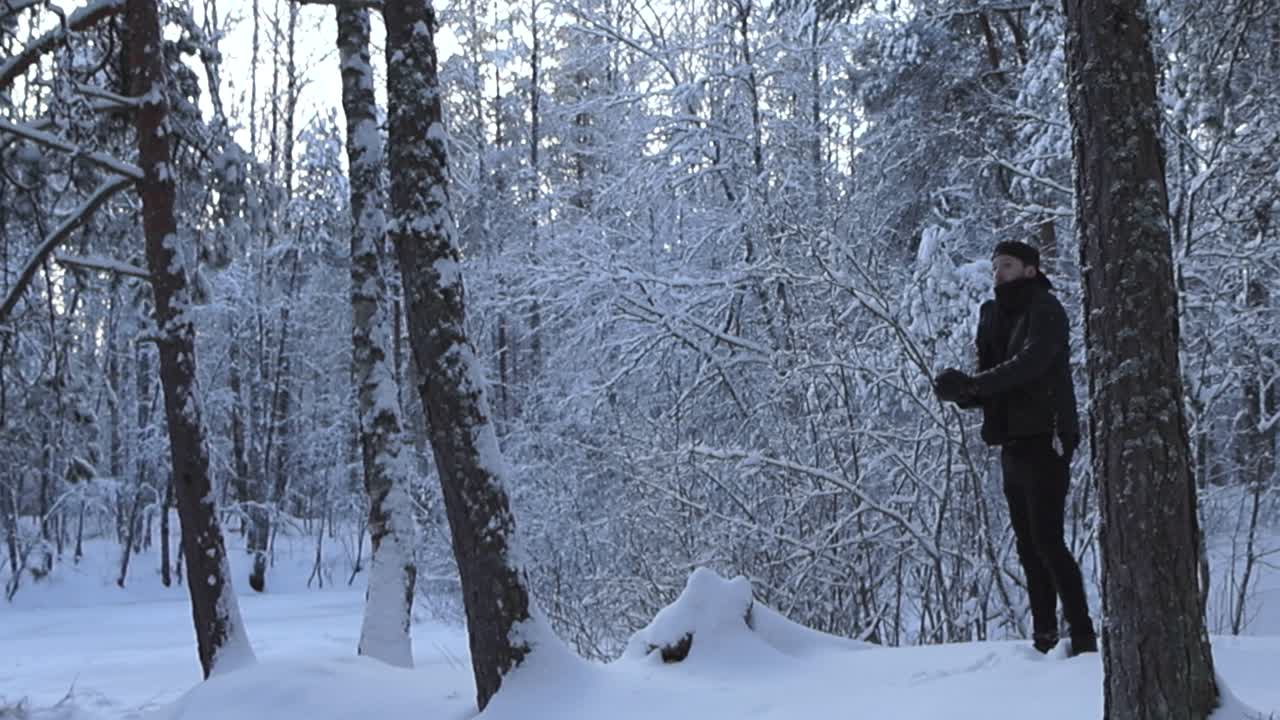 Man with black clothing making a snowball in a winter white dense snow covered forest and throwing the ball against a snow covered tree in slow motion, making the snowball explode and snow flying.