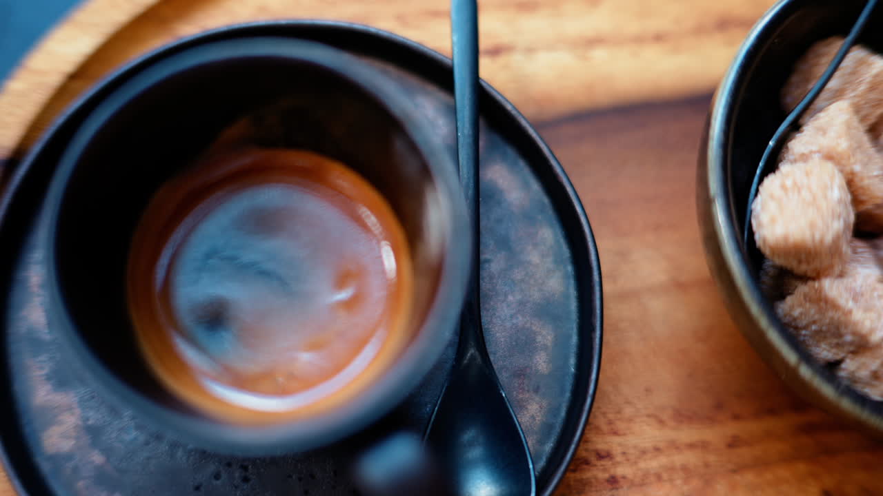 A black cup of espresso with a bowl of brown sugar cubes on a wooden tray