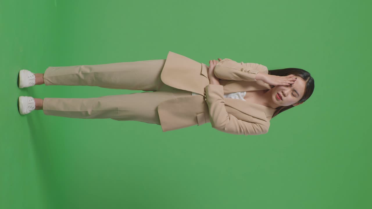 Ganzkörperaufnahme einer asiatischen Geschäftsfrau mit Kopfschmerzen, während sie vor einem Greenscreen steht.