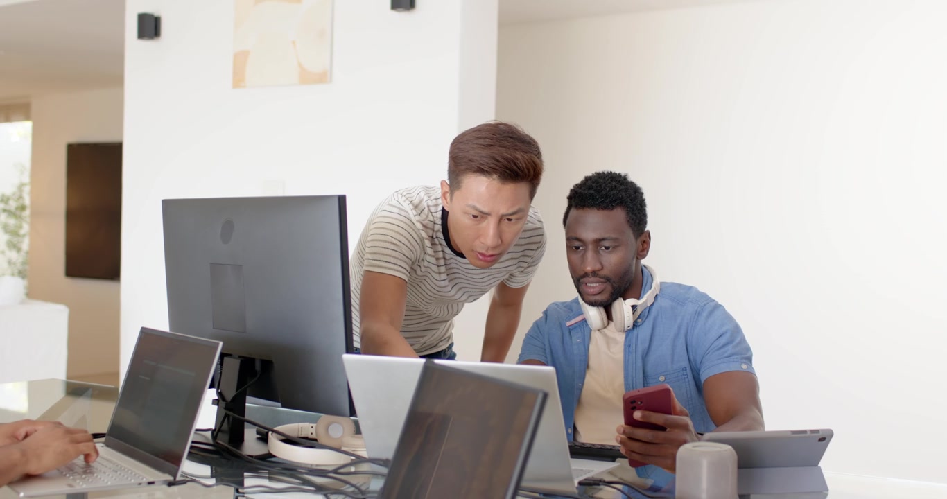 Diverse male coworkers working, showing red phone, leaning over laptops and monitor at glass table