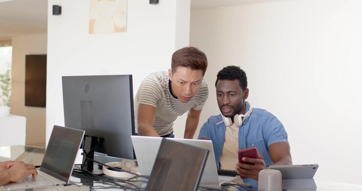 Diverse male coworkers working, showing red phone, leaning over laptops and monitor at glass table