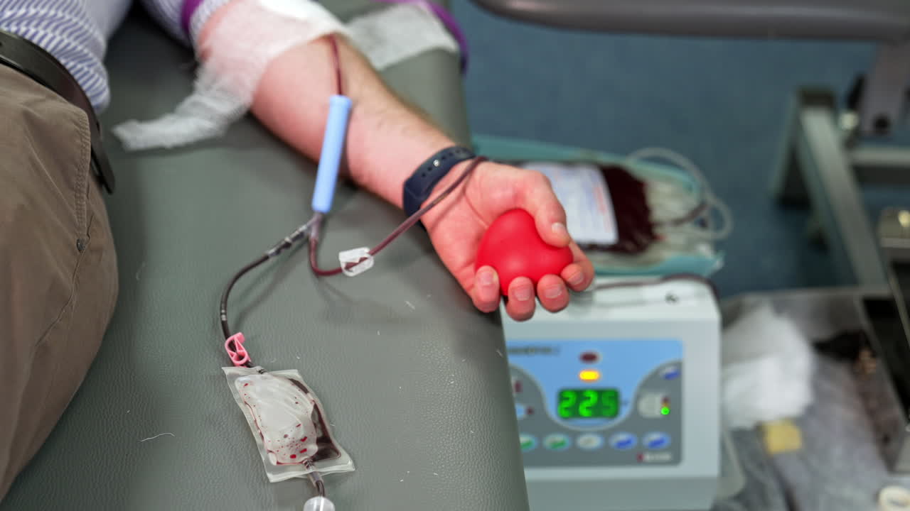 Hand of a male donor holding red rubber heart. Man donating his blood in the hospital. Close up.