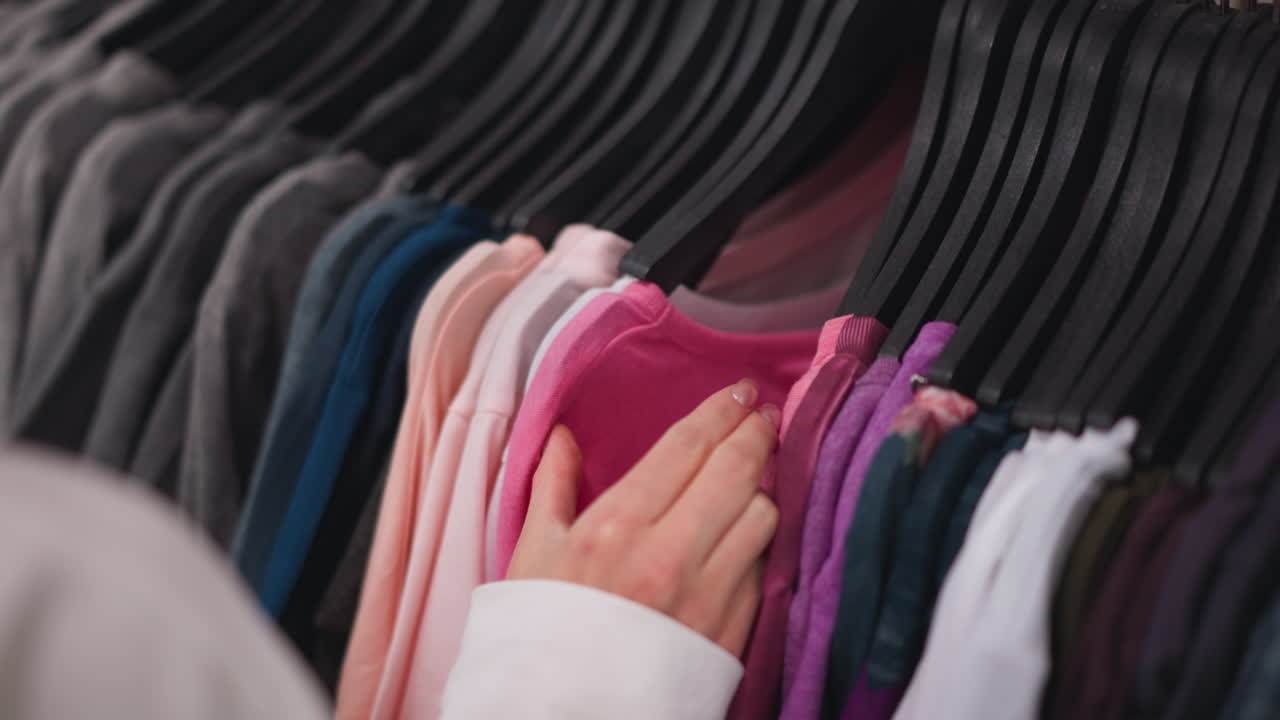 Close-up of female hand browsing through neatly arranged clothing rack, focusing on vibrant pink garment among various colors in a modern retail store
