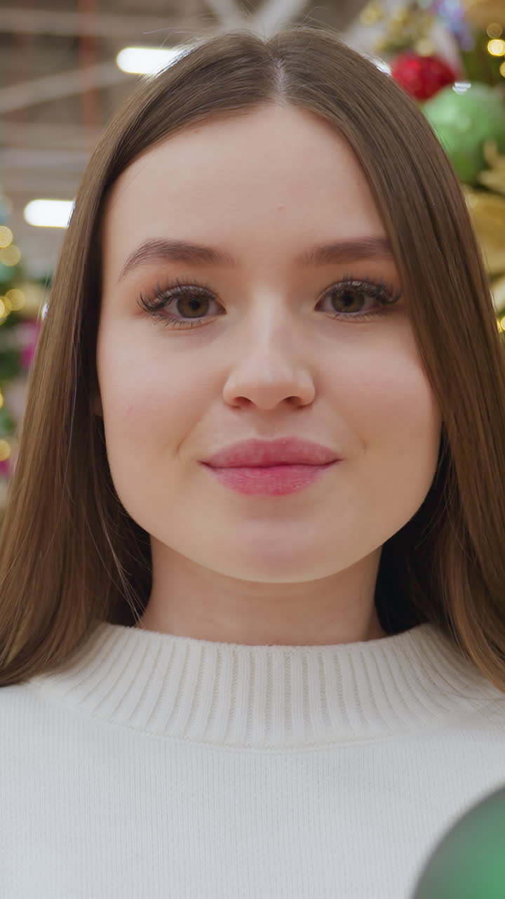 Woman in white sweater holds two green Christmas ornaments, admiring them in a beautiful decor store, the background features a festive Christmas tree and colorful holiday decorations
