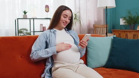 smiling pregnant woman using smartphone text messaging social media chatting sitting on home sofa