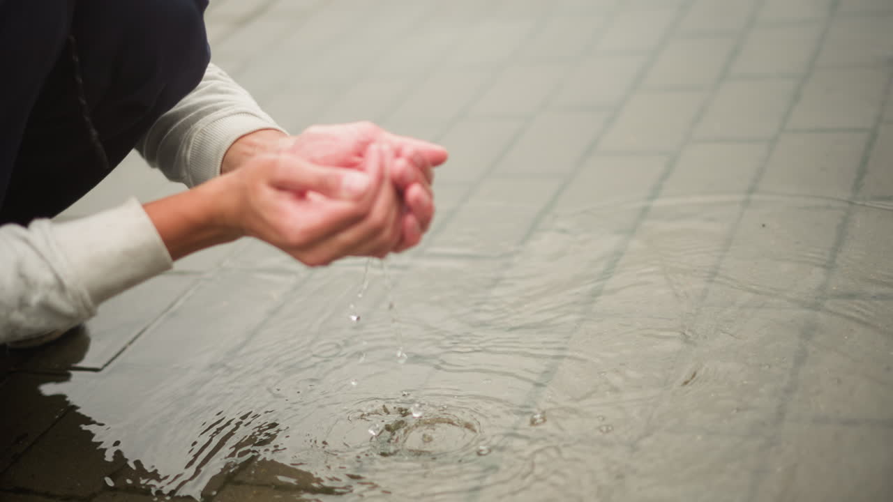 젖은 포장 도로에 웅크리고 앉아 웅덩이에서 빗물을 퍼 올리는 아시아 남성, 타일 표면을 스치는 손, 물이 튀고 물결이 이는 디테일, 젖은 머리카락과 물이 뚝뚝 떨어지는 안경, 자연스러운 거리 풍경을 담은 사진