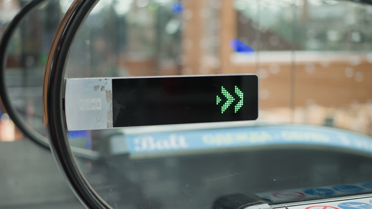 side view glass mounted green arrow led display pointing downward at entrance of moving walkway in mall, blurred silhouettes of shoppers reflected on glass panel under bright overhead lights