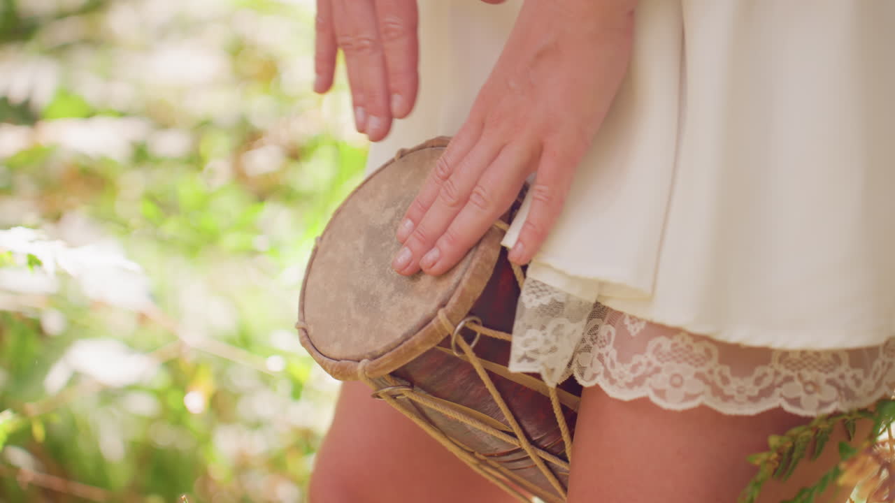 Close up side view of lady in short white gown beating traditional hand drum outdoors, sunlight filtering through green foliage creating warm natural atmosphere of rhythm, culture, and serenity
